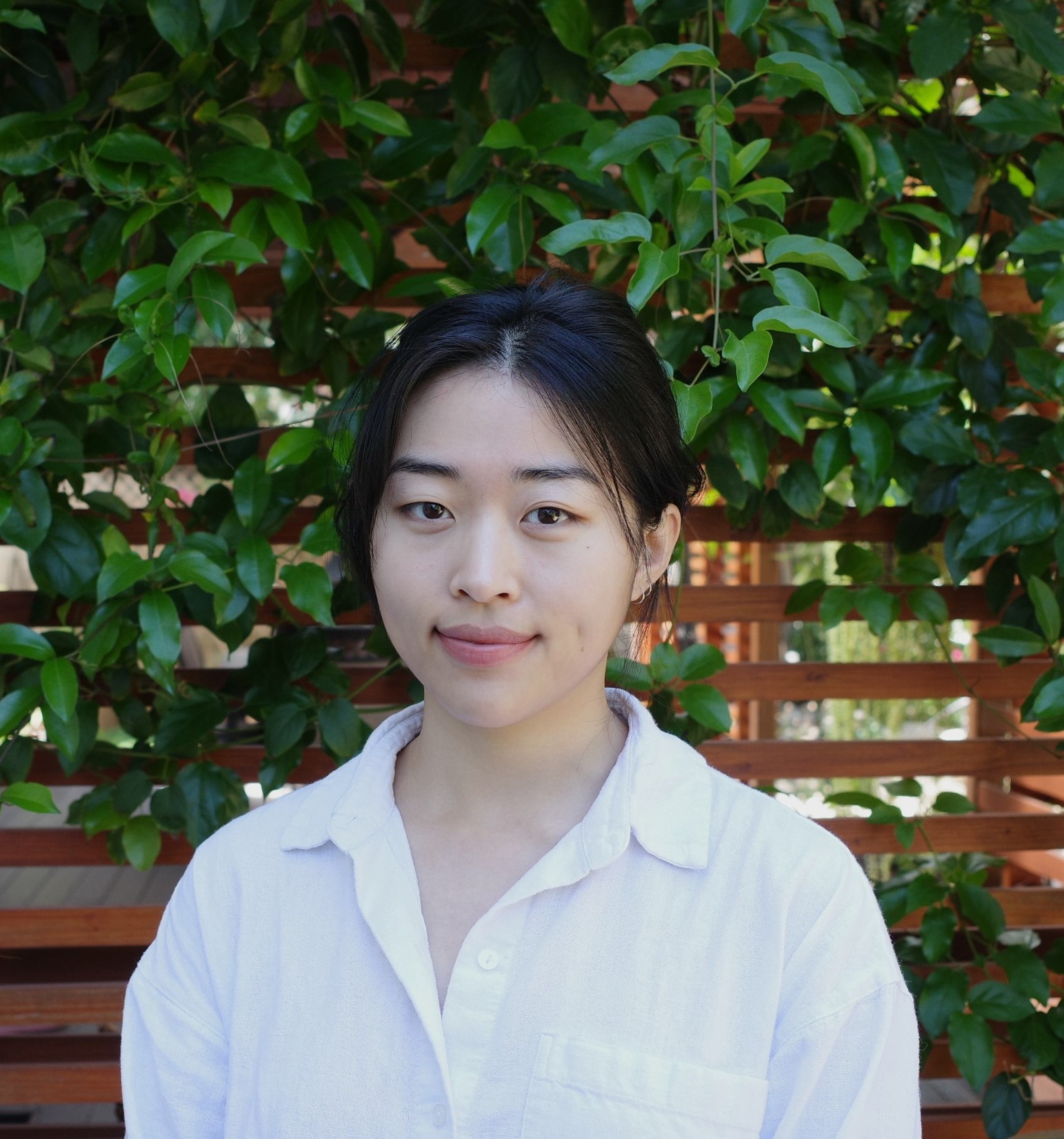 image of sohyeon standing in front of a garden wall covered in vines. it's bright and sunny, and she is smiling slightly. she is wearing a white button-down and her hair is tied up in a bun.